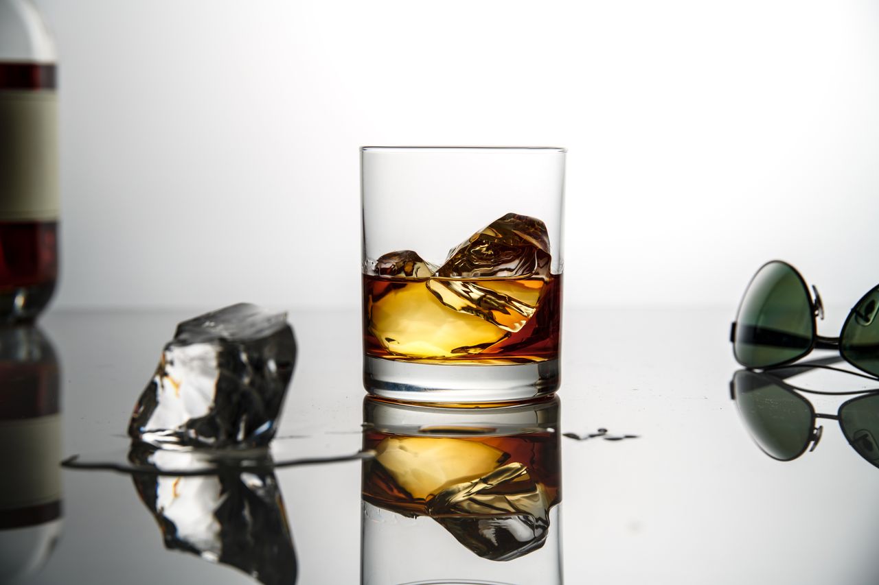 A sophisticated glass of whiskey with ice cubes on a reflective surface, set against a white background.