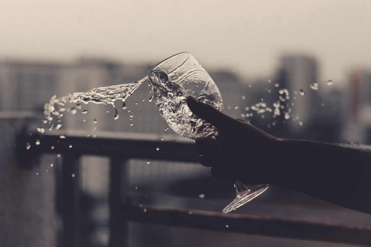 Unrecognizable person pouring water from glass while standing near metal fence on street with blurred buildings on background during daytime